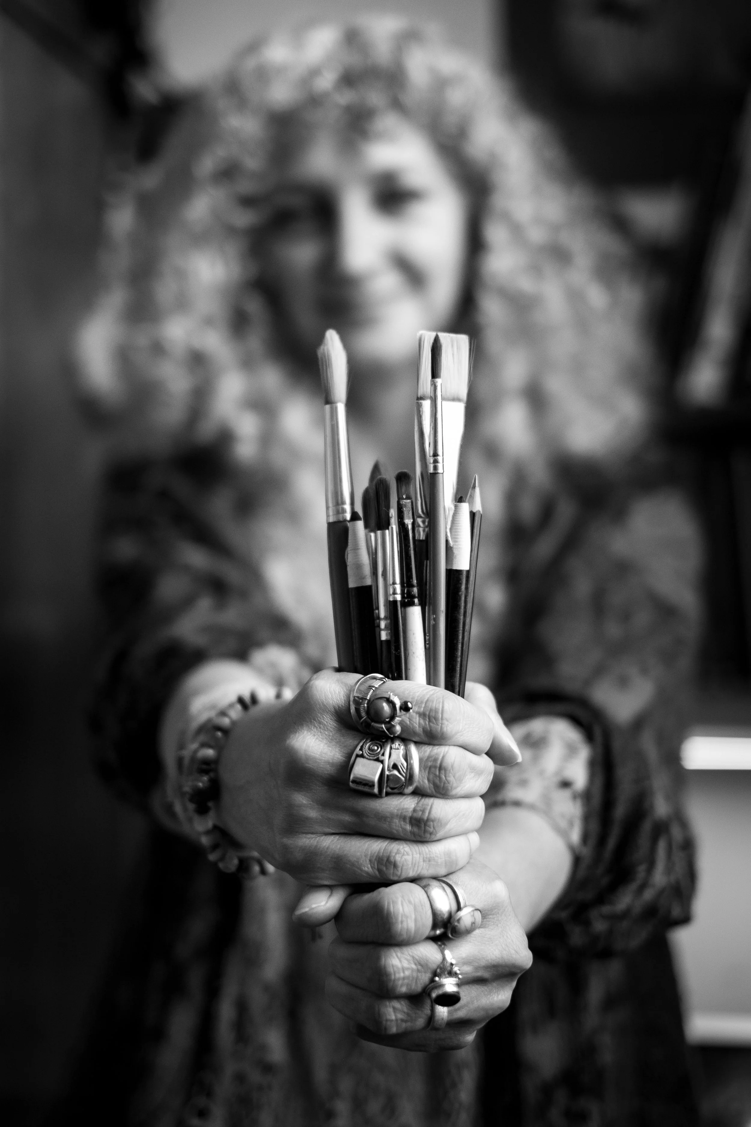 A black & white photo of a woman with long curly hair holding out infant of her a collection of artists brushes