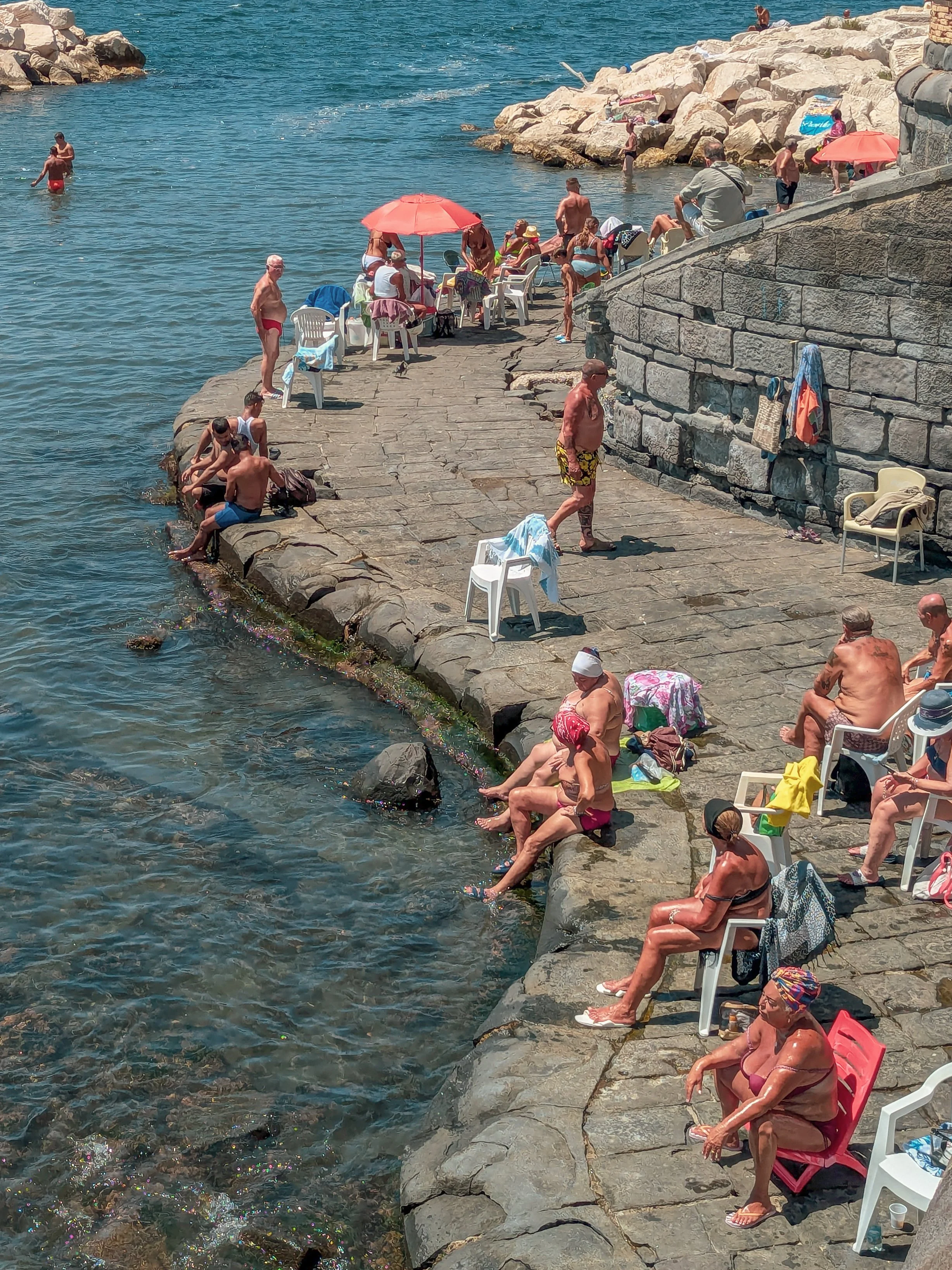 People relaxing on a rocky shoreline and swimming in the water on a sunny day, with some sitting on chairs, some under umbrellas, and others in the water.