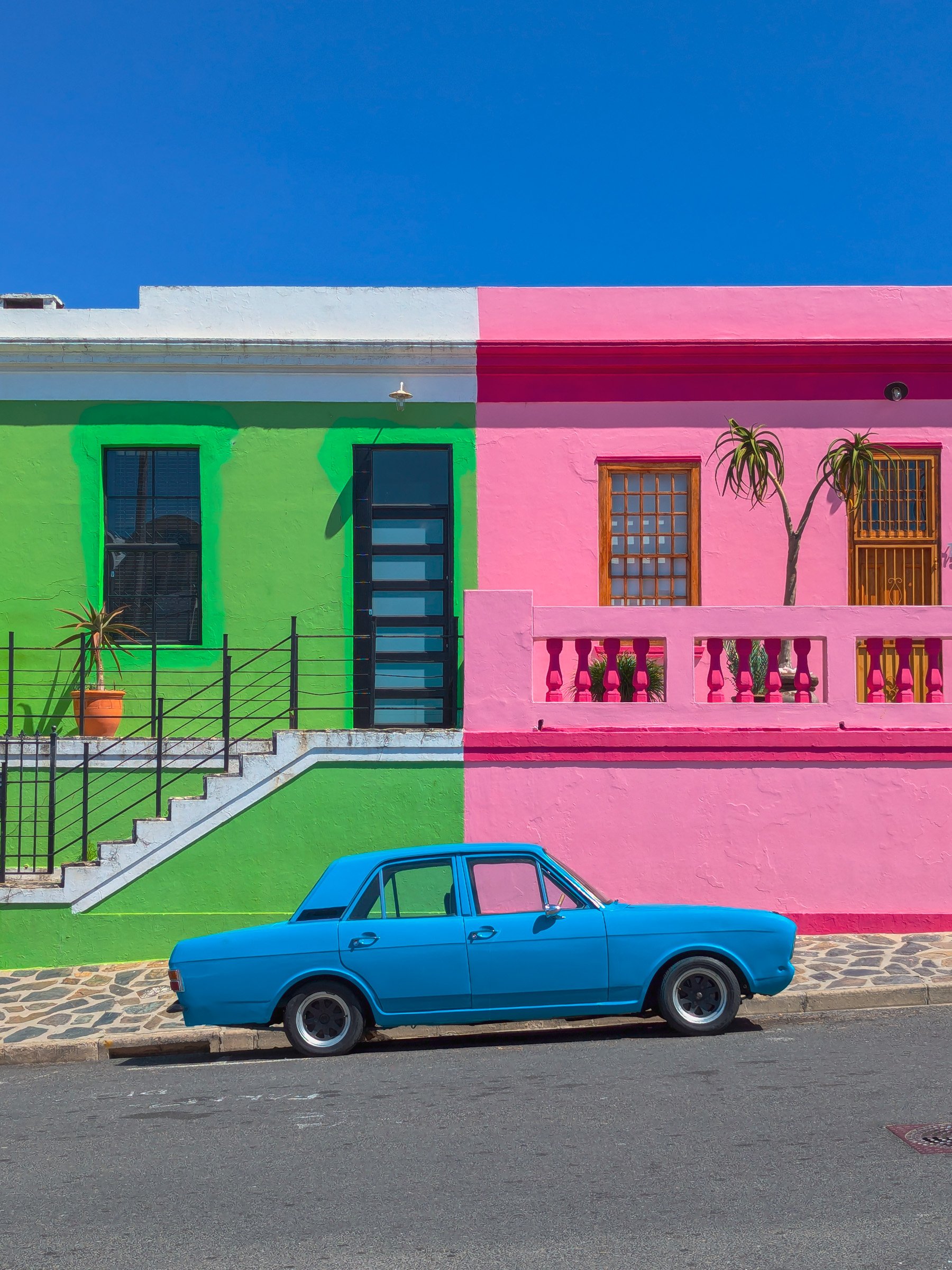 Colorful, split-color building with green on the left and pink on the right, featuring two windows and a door with stairs and potted plants; a bright blue vintage car parked in front under a clear blue sky.