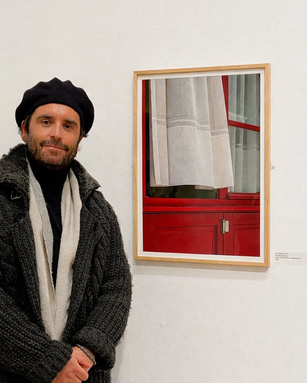 A man with a beard and dark hair wearing a black beret, dark sweater, and gray scarf standing next to a framed photograph hanging on a white gallery wall.