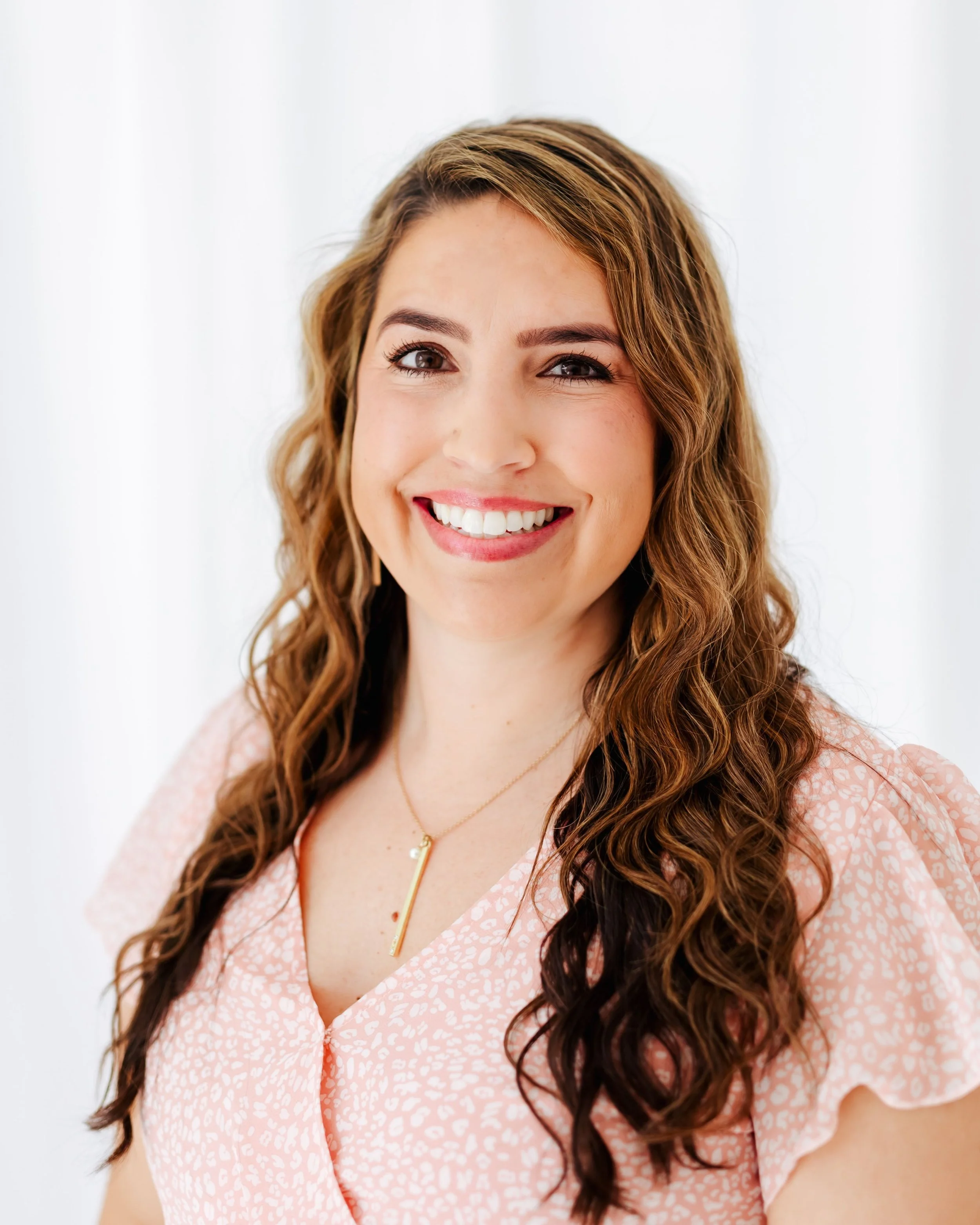 A woman with long wavy brown hair smiling while looking at the camera, wearing a pink patterned blouse and a delicate gold necklace against a white background.