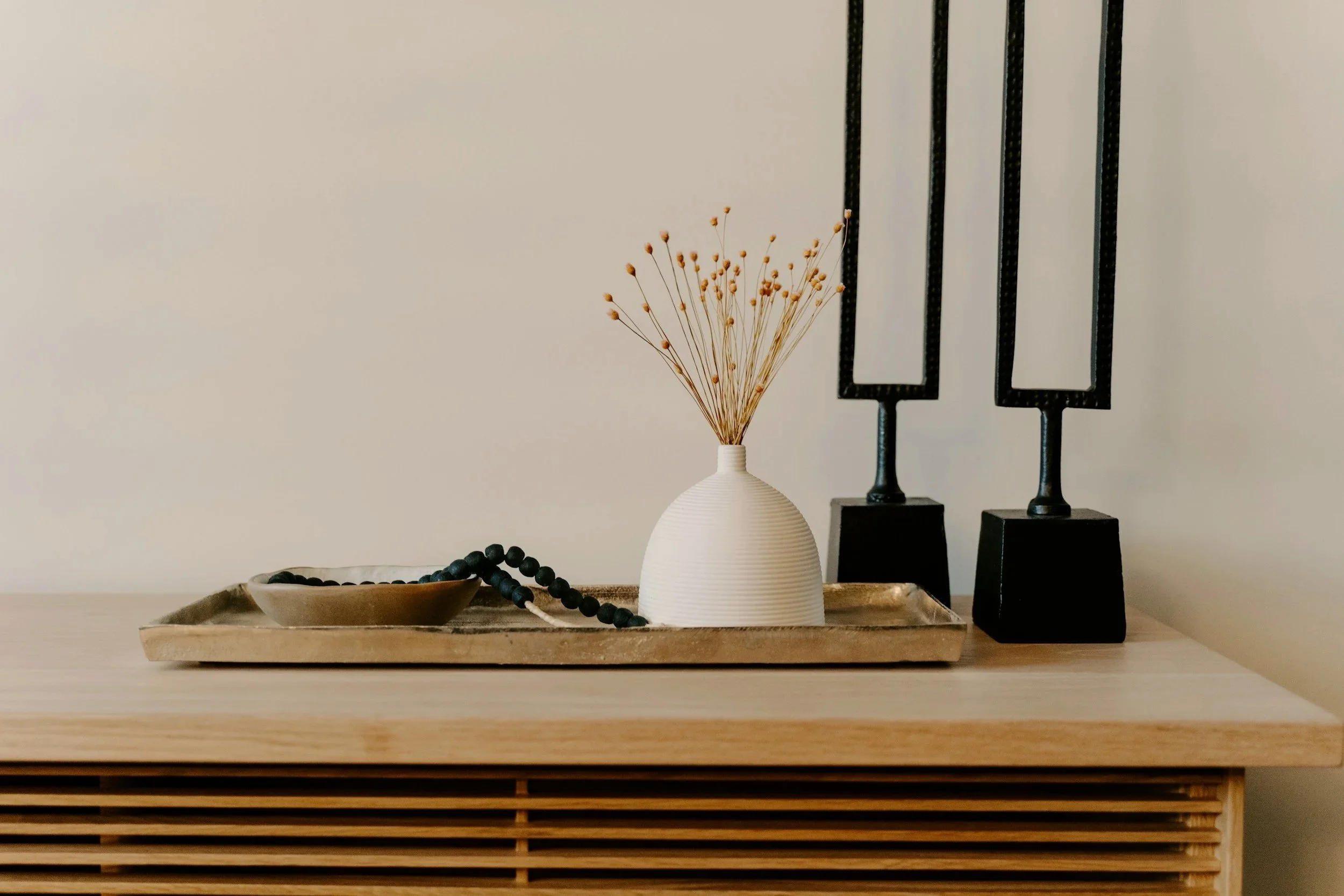 A beige vase with dried flowers on a wooden tray. Next to the vase is a black bead necklace, and behind it there are two black candle holders with rectangular openings, all on a wooden surface against a plain white wall.