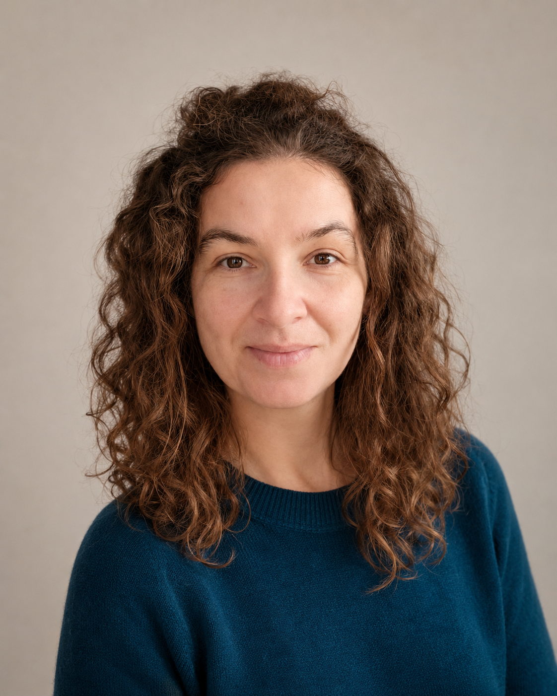 A woman with curly brown hair wearing a blue sweater, looking at the camera with a neutral expression against a plain beige background.