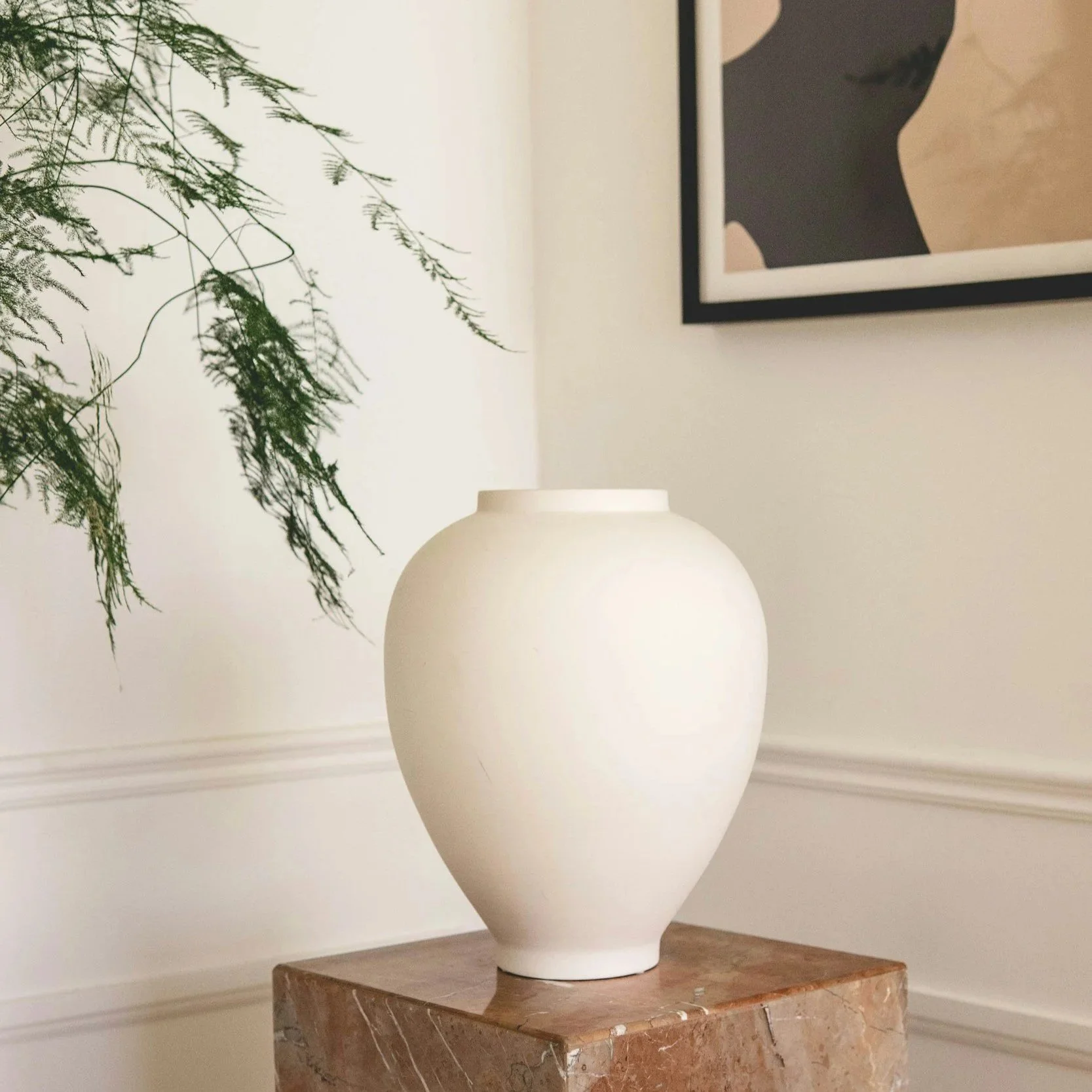 A large white ceramic vase with a smooth surface and rounded shape, placed on a brown marble pedestal in a room with off-white walls. Part of a framed black-and-white artwork is visible on the wall, and some green leafy foliage extends into the frame from the left side.