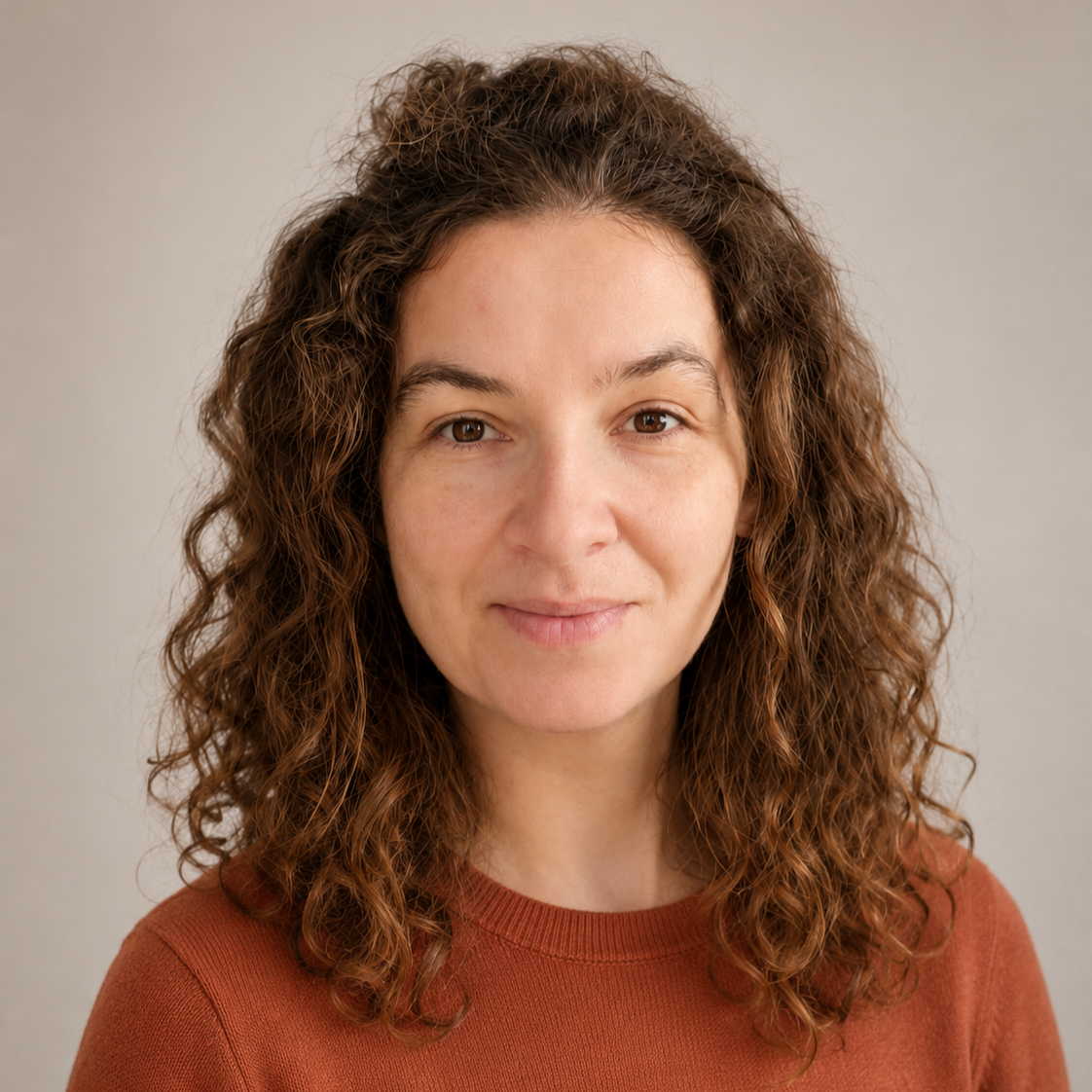 Close-up portrait of a woman with curly brown hair wearing a rust-colored top, against a neutral background.