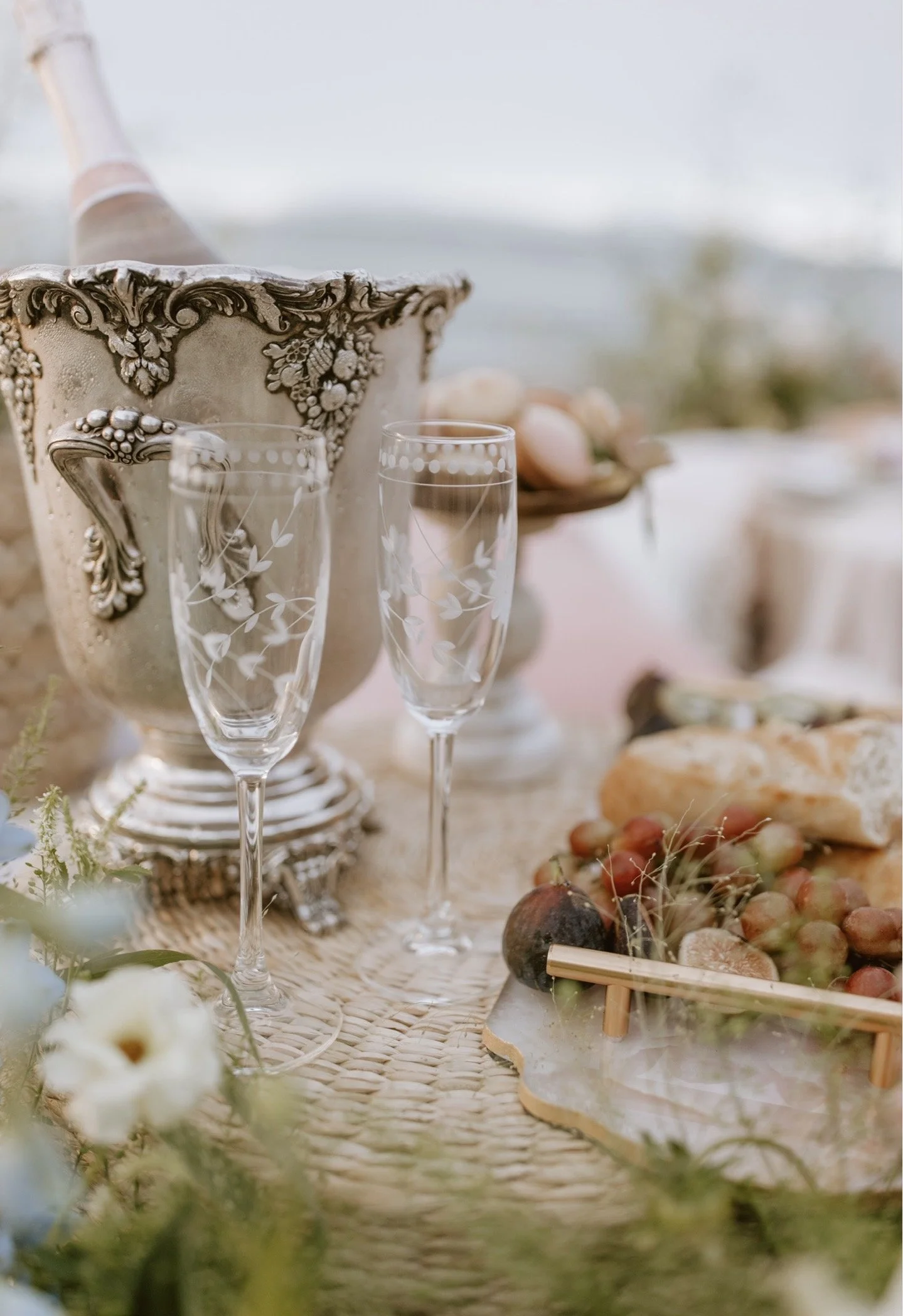 A picnic setup on a grassy lawn with a white umbrella, flowers, candles, and a wooden table, with lush greenery and historic architecture in the background.
