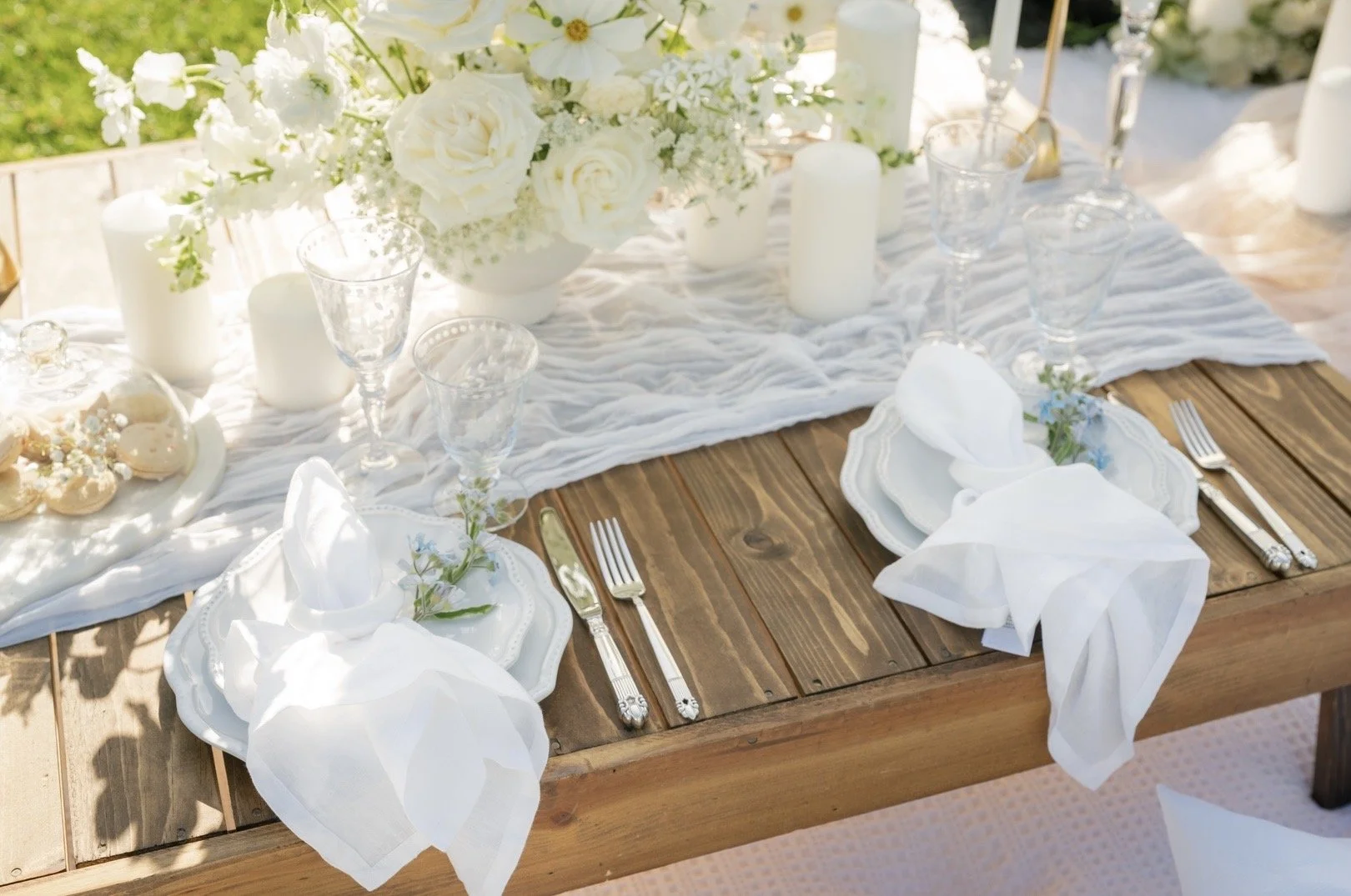 Elegant outdoor dining table set with white plates, napkins, silverware, glassware, and a flower arrangement of white flowers, surrounded by candles, on a wooden table with a white table runner.