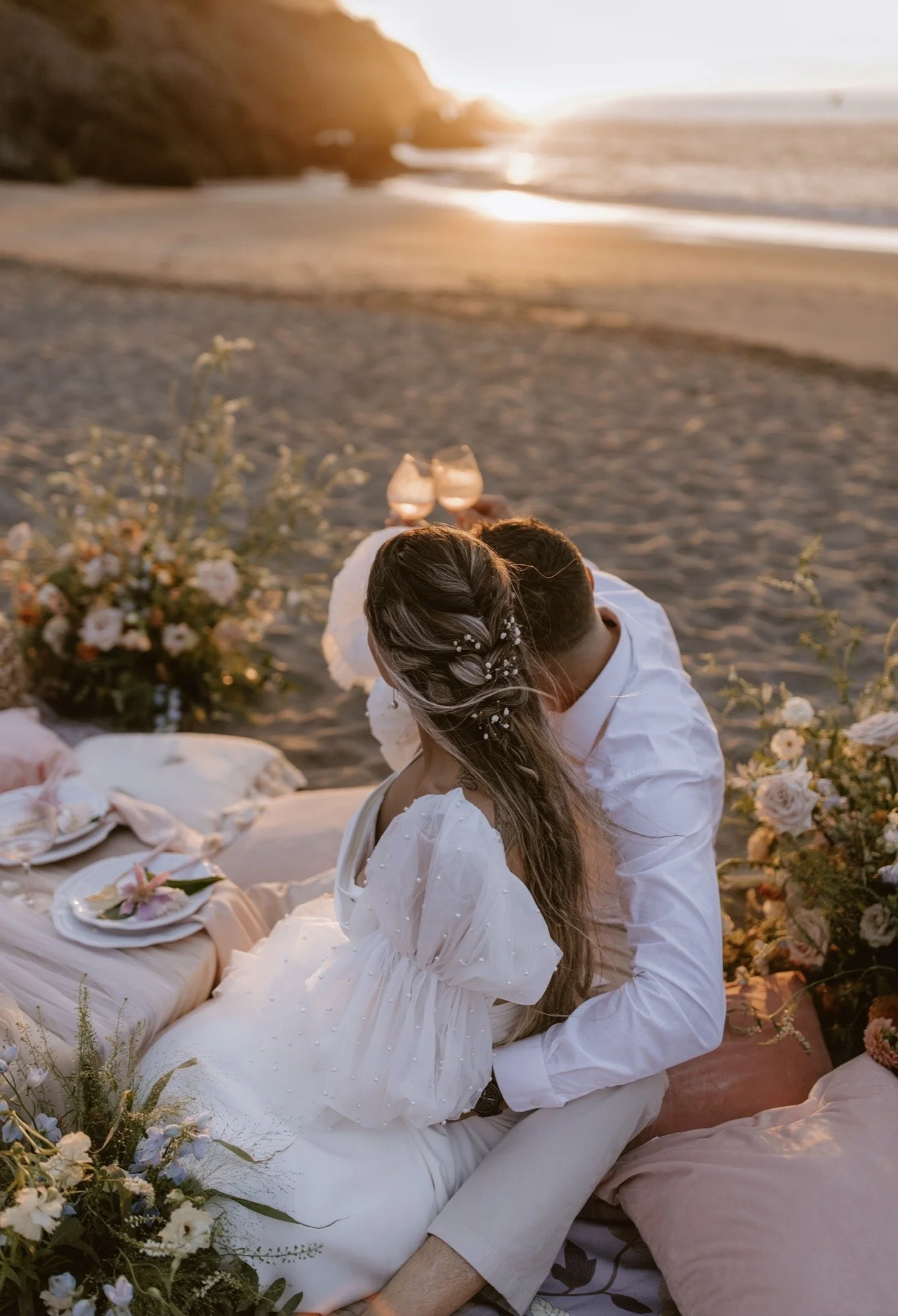 Elegant outdoor dining table decorated with pink, purple, and white flowers, pink candles, and lace tablecloth, set in a garden with lush greenery.