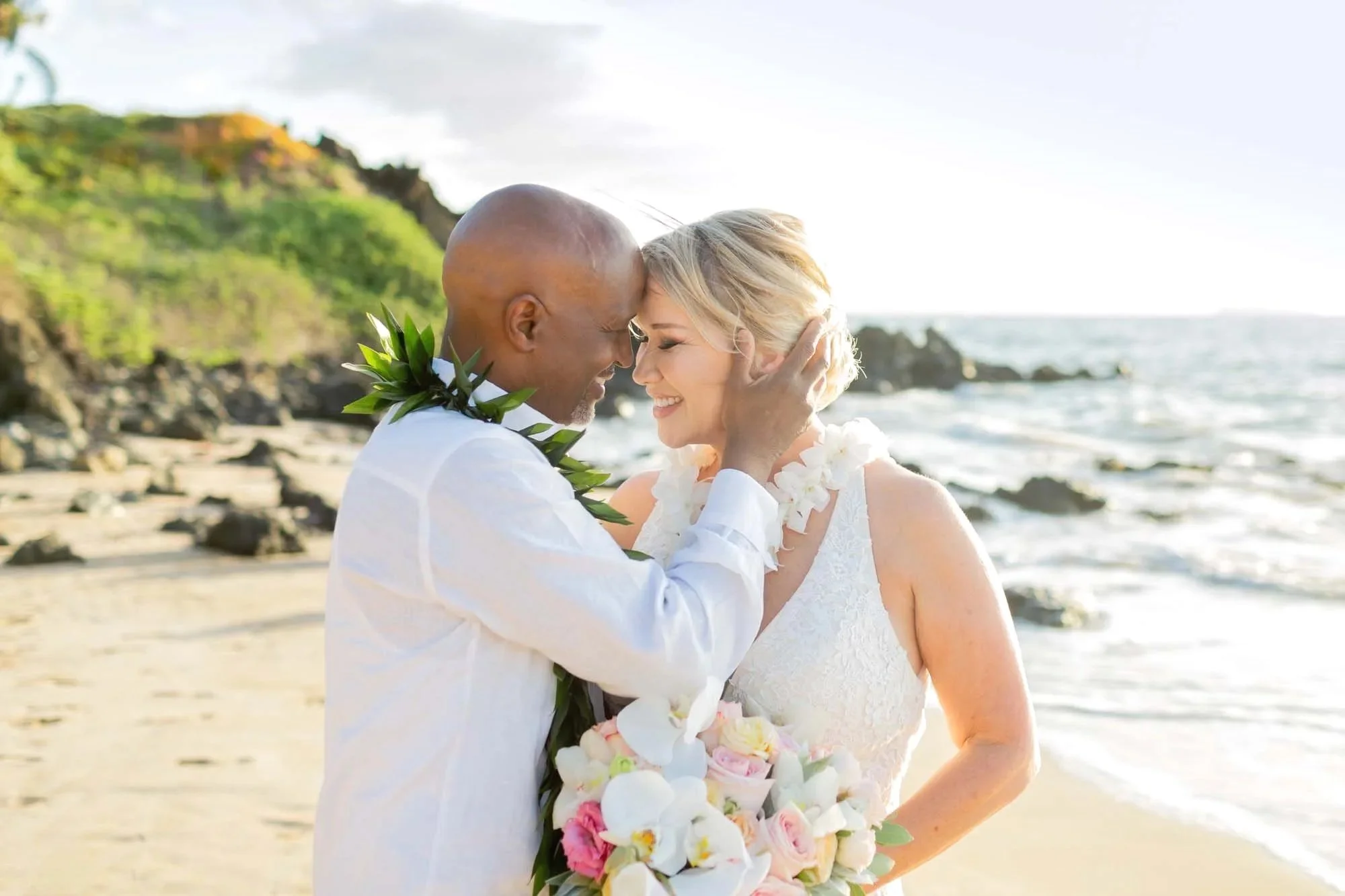 A bride and groom sharing a kiss on the beach, dressed in wedding attire, with the ocean and greenery in the background.