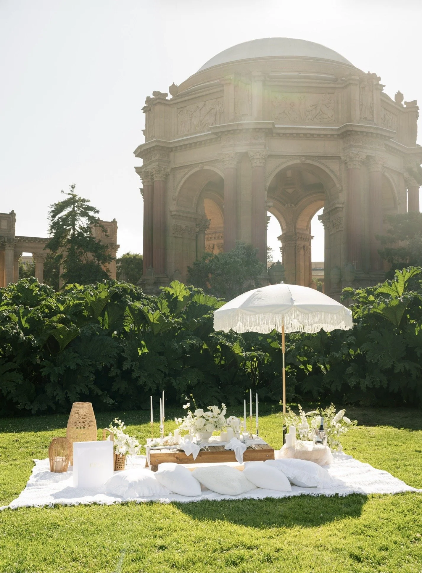 An outdoor wedding setup with a white canopy, decorated table, candles, and white pillows on the grass, with historical architecture and lush greenery in the background.