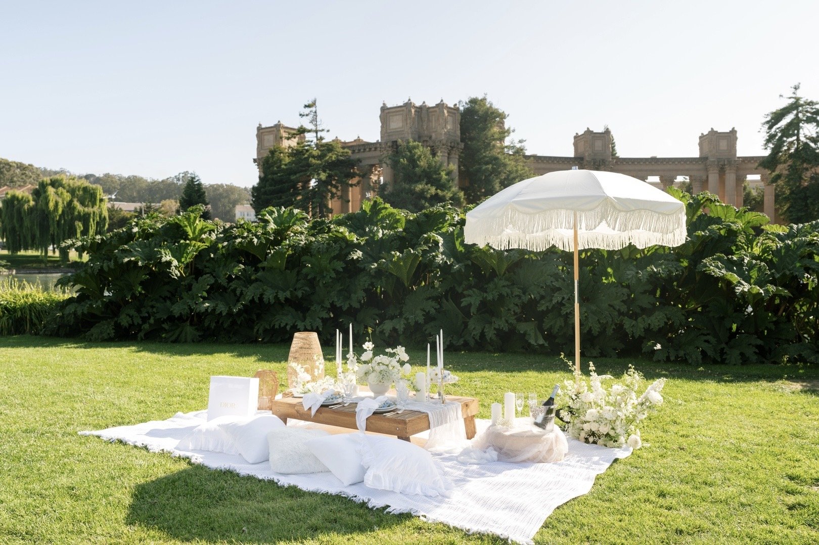 A picnic setup on a grassy lawn with a white umbrella, flowers, candles, and a wooden table, with lush greenery and historic architecture in the background.