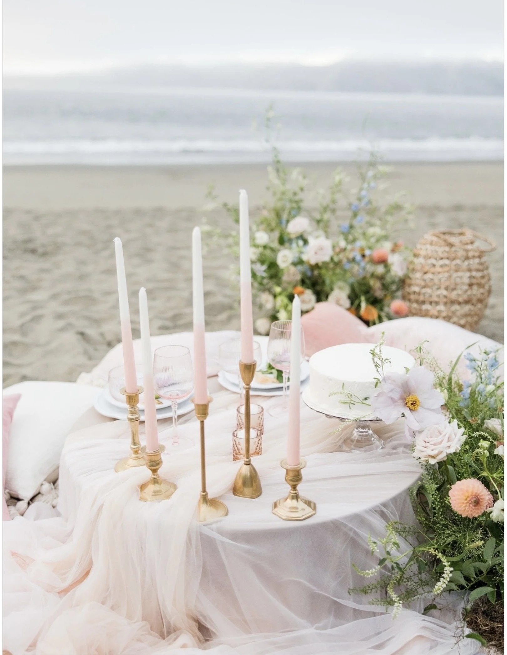 Beachside table setting with white candles, pink and gold candle holders, floral arrangements, and a cake, overlooking the ocean with cloudy sky.
