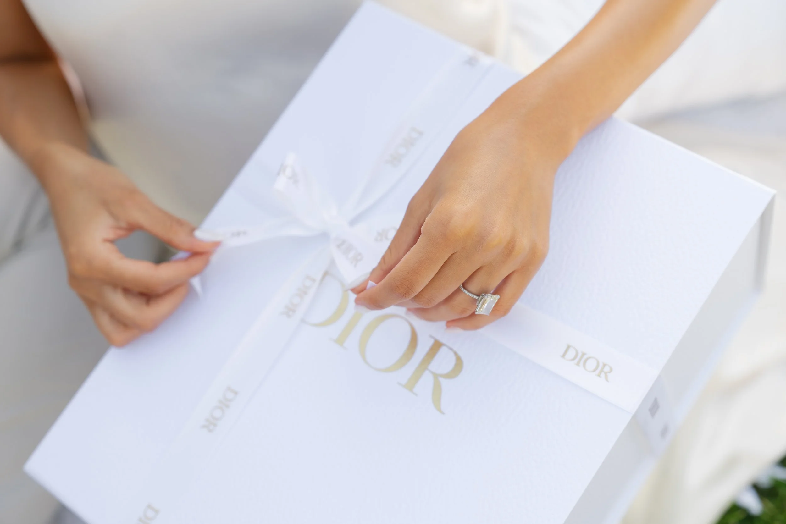 Close-up of a person opening a white Dior box decorated with gold lettering and a ribbon, revealing a white shopping or gift bag inside.