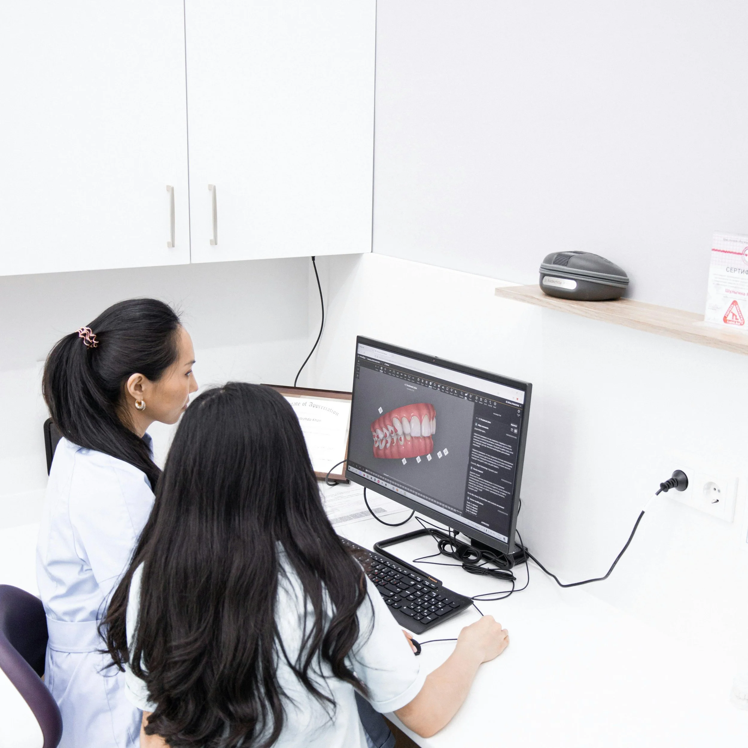 Two women are working together at a computer in a medical or dental office. One woman is wearing a white coat and has dark hair tied back. They are looking at a monitor displaying dental or medical information, with a model of teeth on the screen.