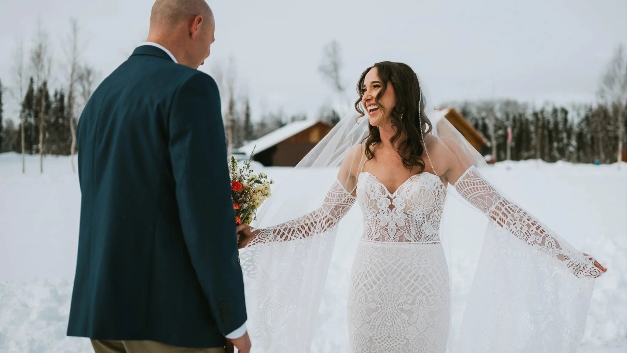 A bride and groom standing outside in a snowy landscape, holding hands and smiling at each other during their wedding ceremony.