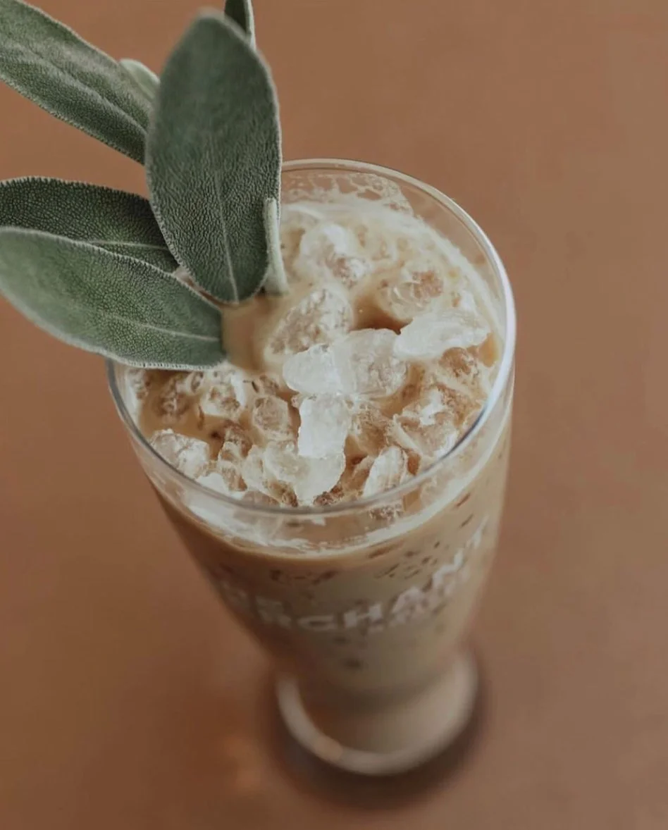 A tall glass filled with iced coffee or latte, topped with ice cubes and garnished with fresh sage leaves.