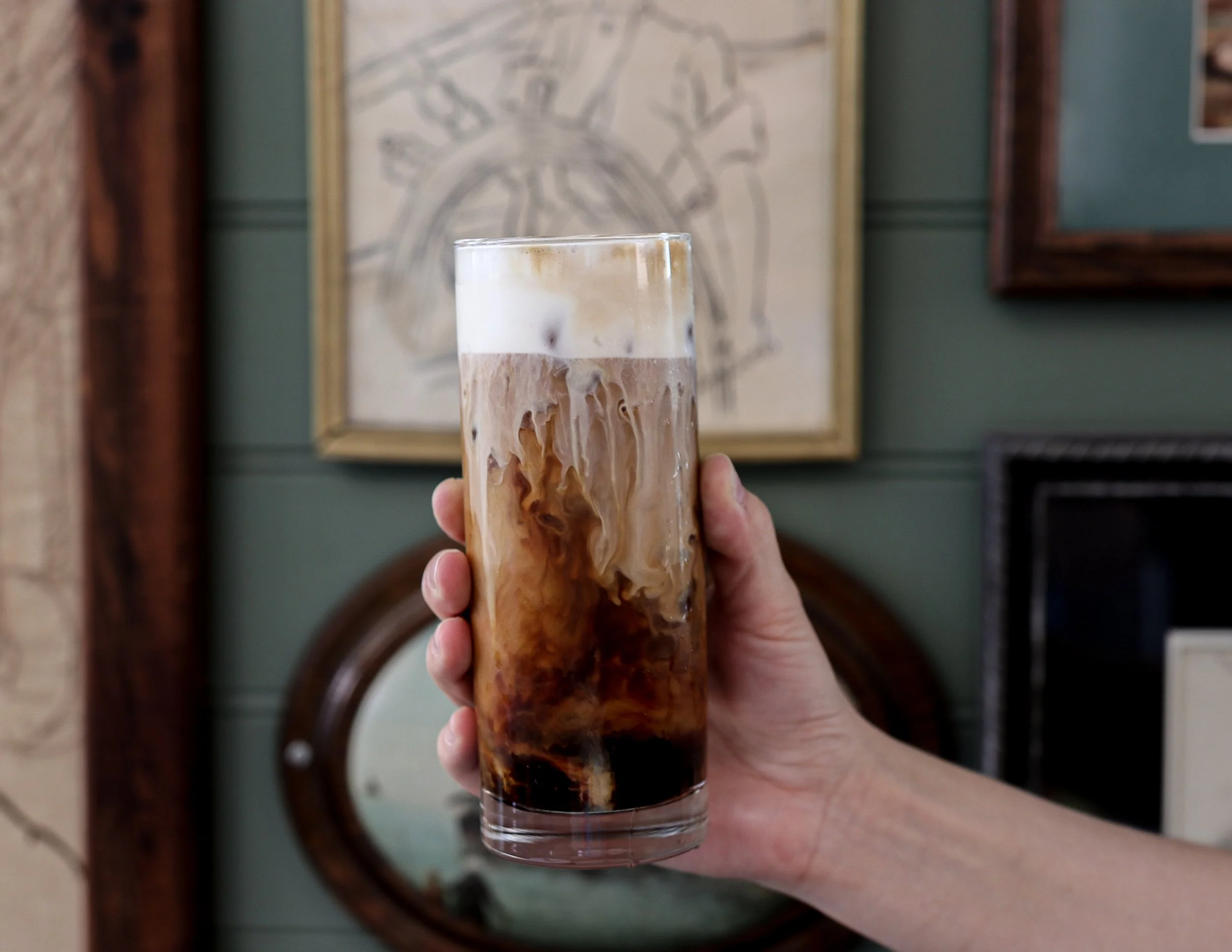 Hand holding an iced latte with cream swirling into the coffee