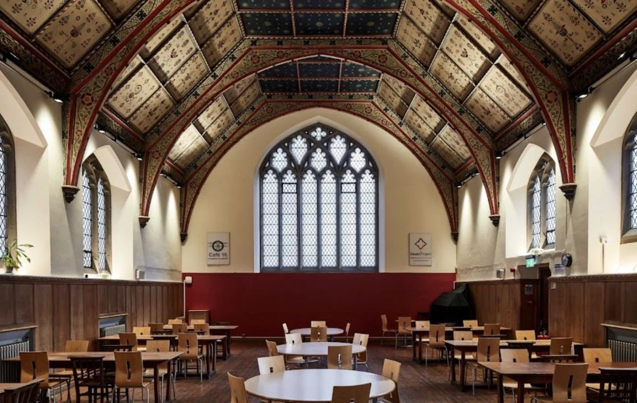 Interior of a historic building with high, ornate, vaulted ceiling, stained glass windows, and wooden tables and chairs.
