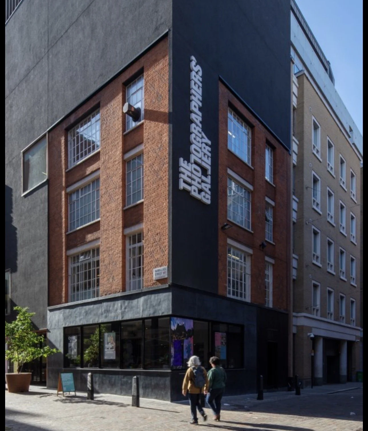 Street view of a multi-story brick building with large windows and a black sign on the corner that reads 'The PG Kapp & Sons Gallery.' Two people walk on the sidewalk in front of the building.