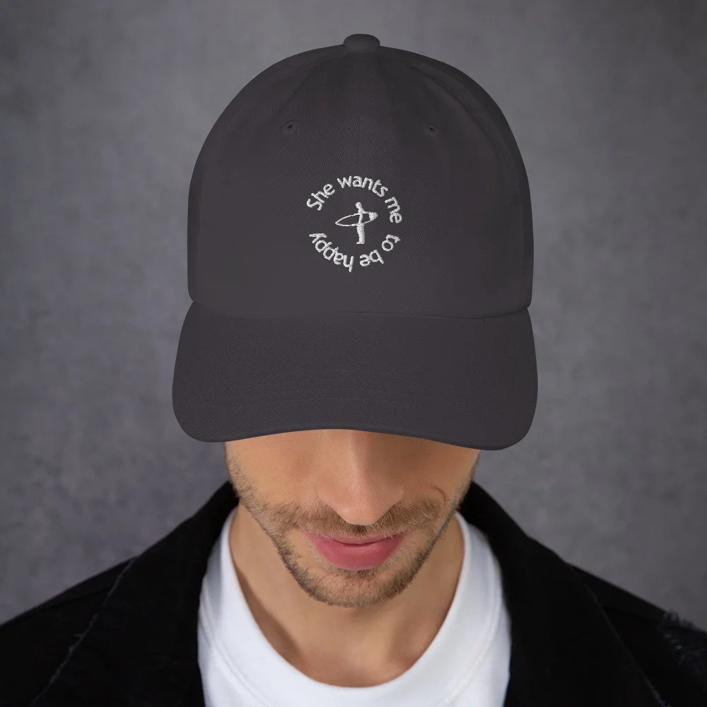 Black baseball cap with embroidered white text and a small graphic on the front, worn by a man with a light beard and mustache, against a gray background.
