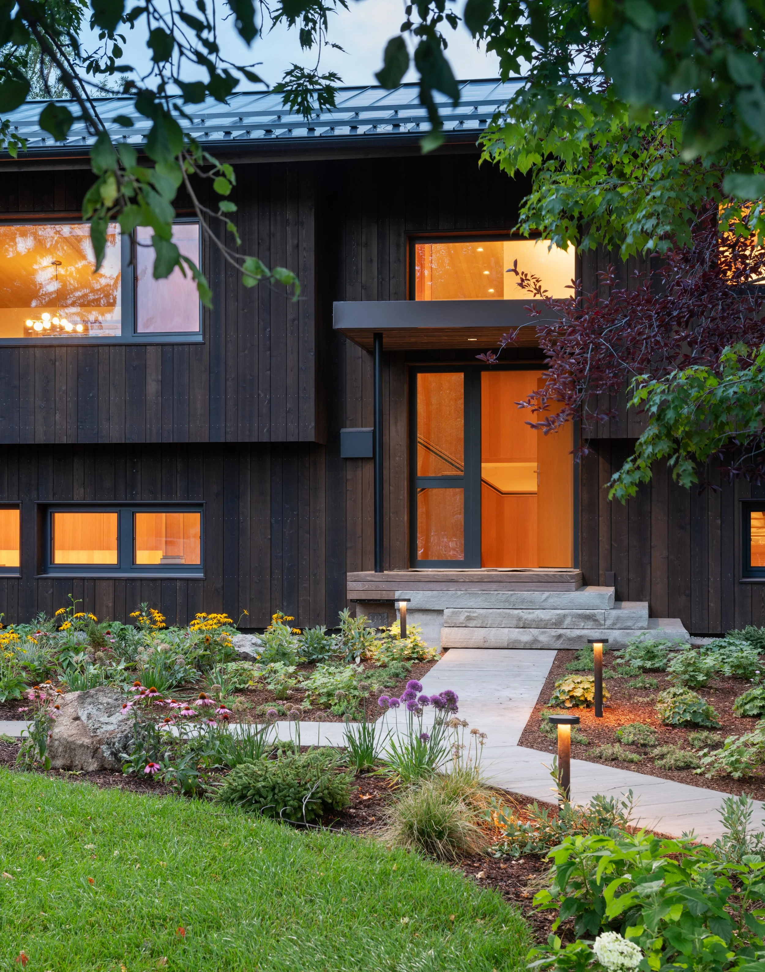 A modern house with dark wooden exterior and large windows, surrounded by a colorful garden with flowers and plants, illuminated by pathway lights, during dusk.