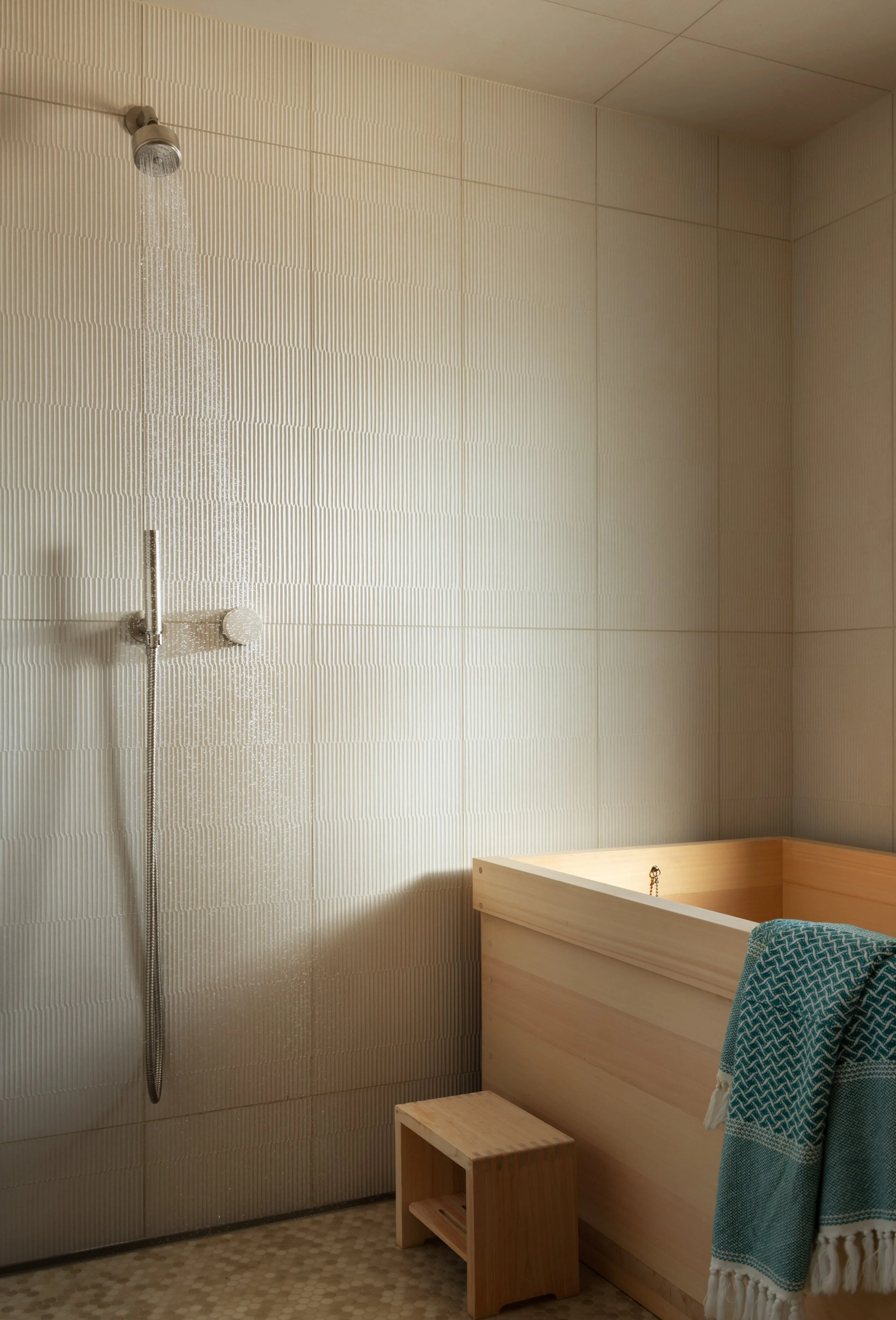 Bathroom with beige textured wall tiles, a shower head, a wooden bathtub, and a small wooden footstool, with a blue towel hanging over the bathtub.