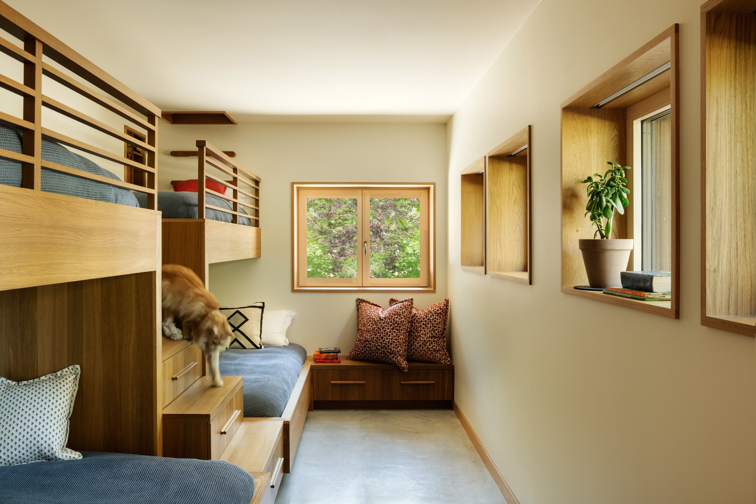 A cozy bedroom with a wooden bunk bed and a small sitting area near a window with greenery outside. There is a dog on the lower bunk, and decorative pillows and books on the seating area.