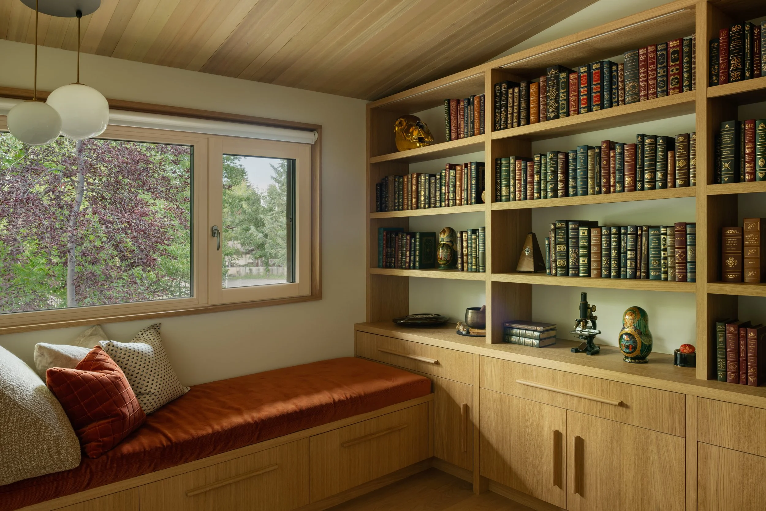 Corner of a room with a large window showing trees outside, a built-in wooden bench with pillows, and a tall wooden bookshelf filled with books and decorative items, under a wooden ceiling with hanging light fixtures.