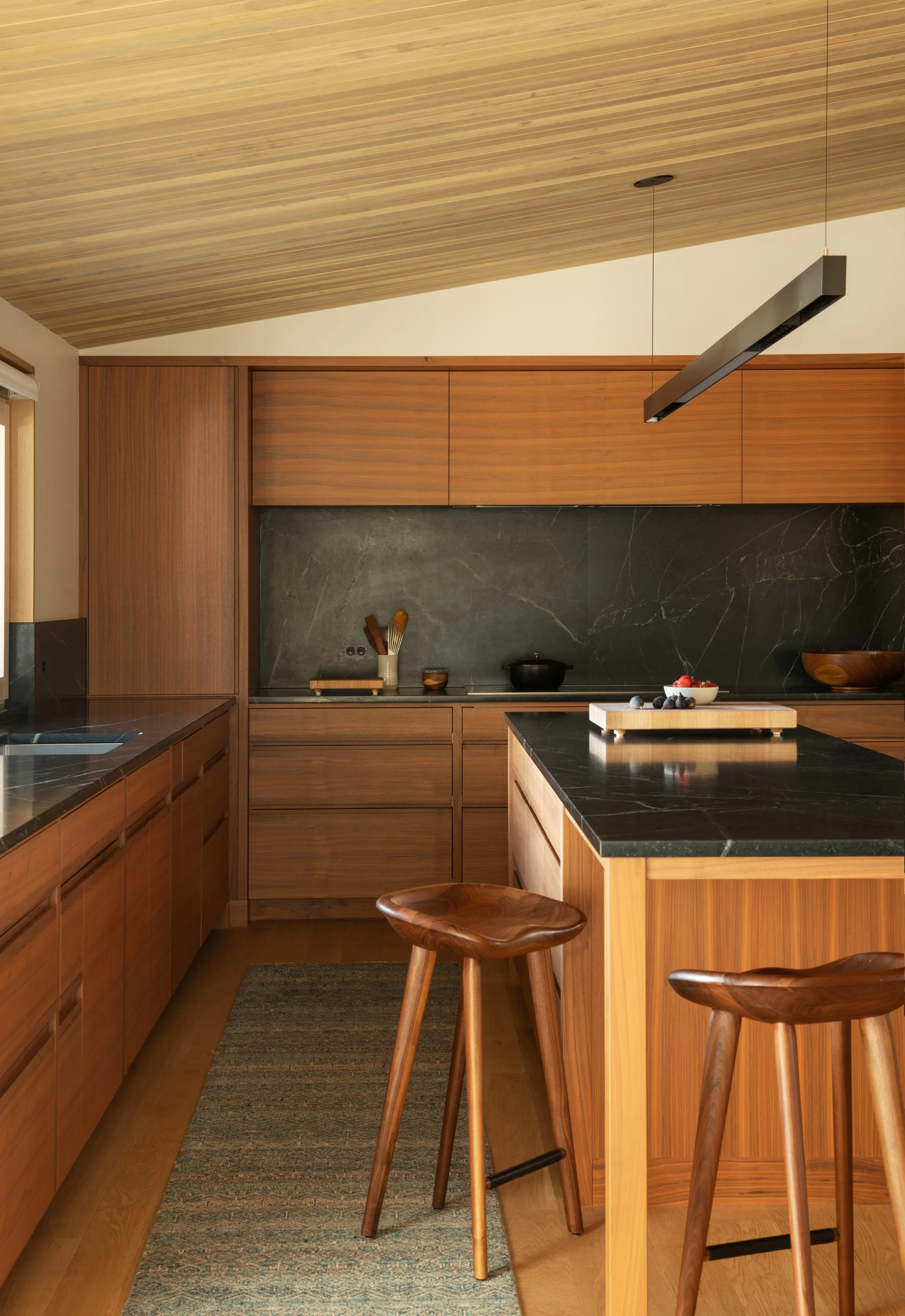 Modern kitchen with wooden cabinets, black marble countertops, a kitchen island with a bowl of fruit, and wooden bar stools.