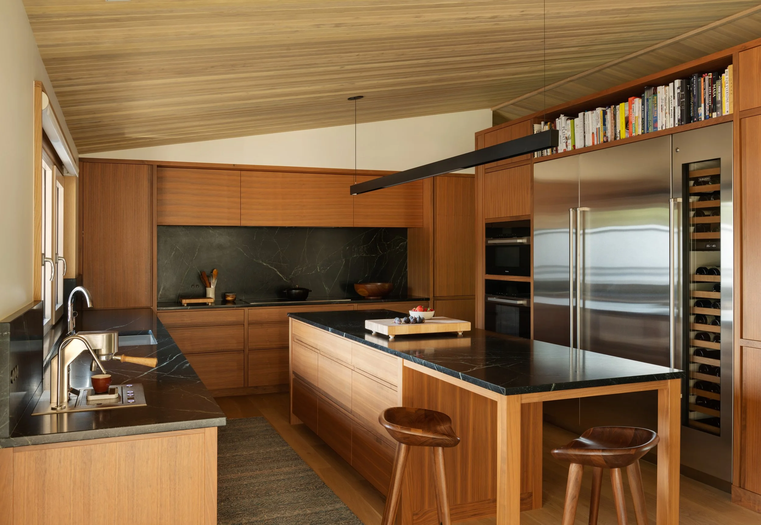 Modern kitchen with wooden cabinets, black marble countertops, stainless steel appliances, and a wood kitchen island with fruit on it.