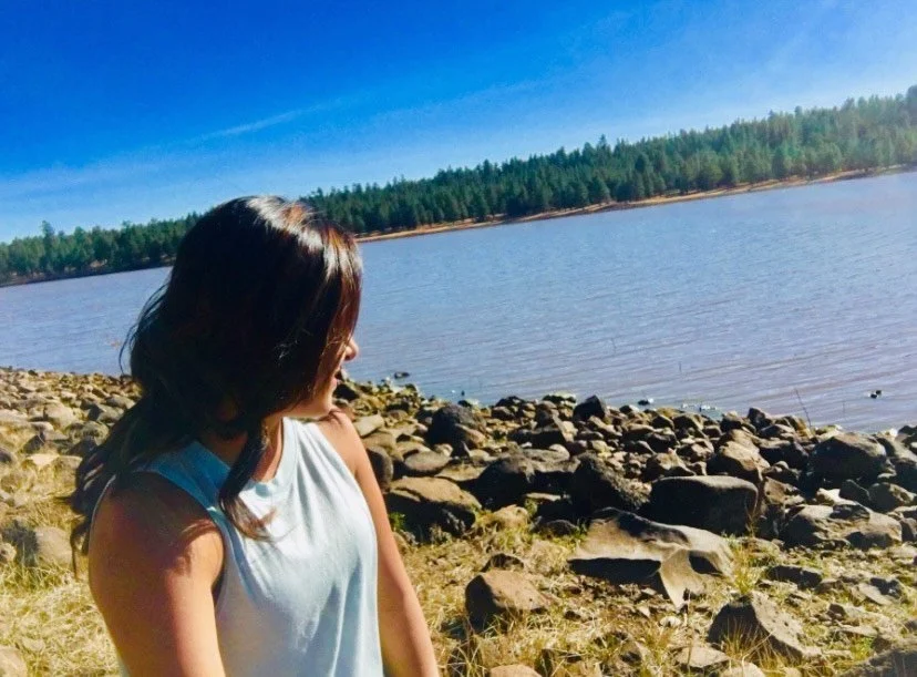 Person near rocky lakeshore with trees and clear blue sky