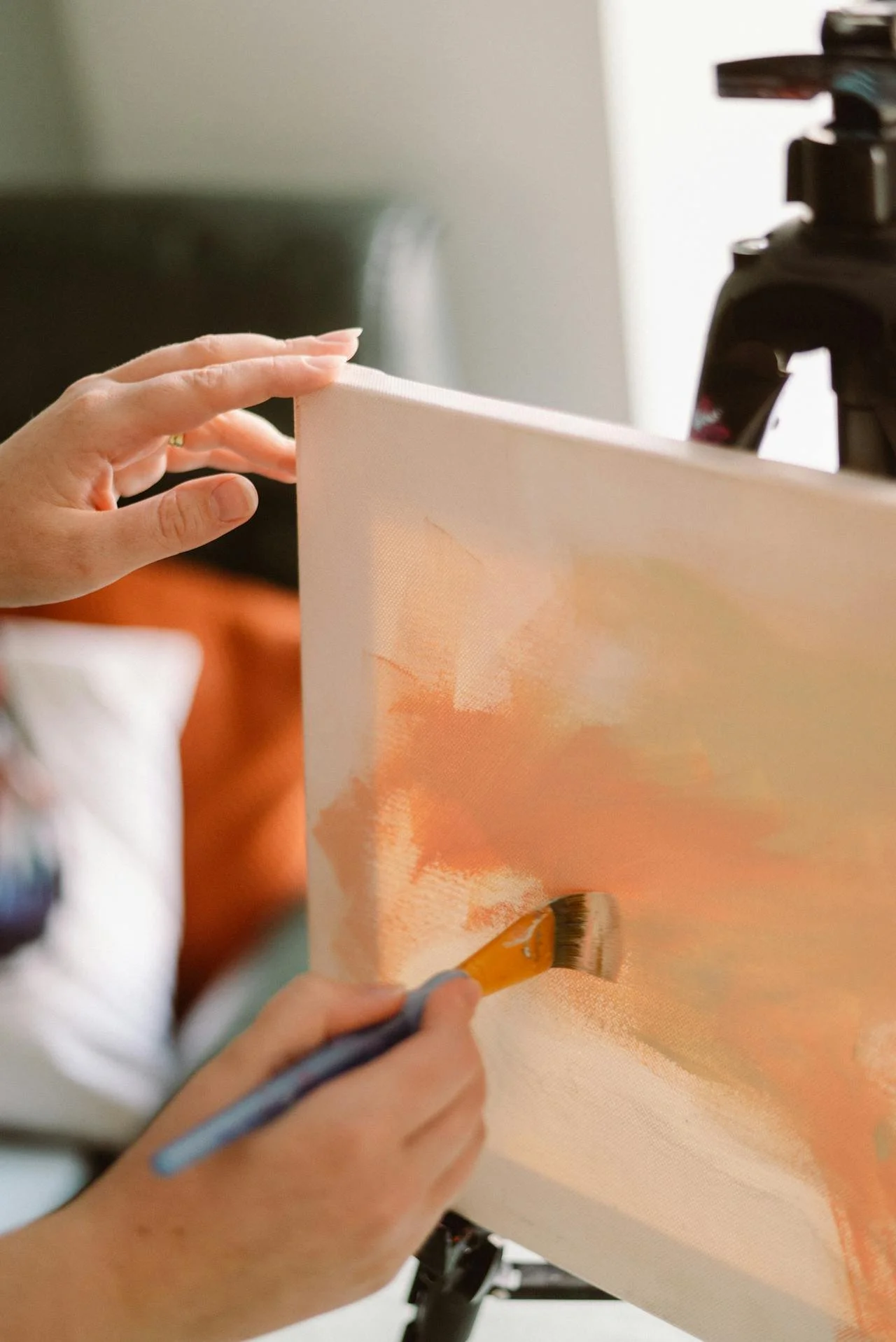 Person painting on a canvas with an orange and white color palette, using a brush; the canvas is held on an easel.