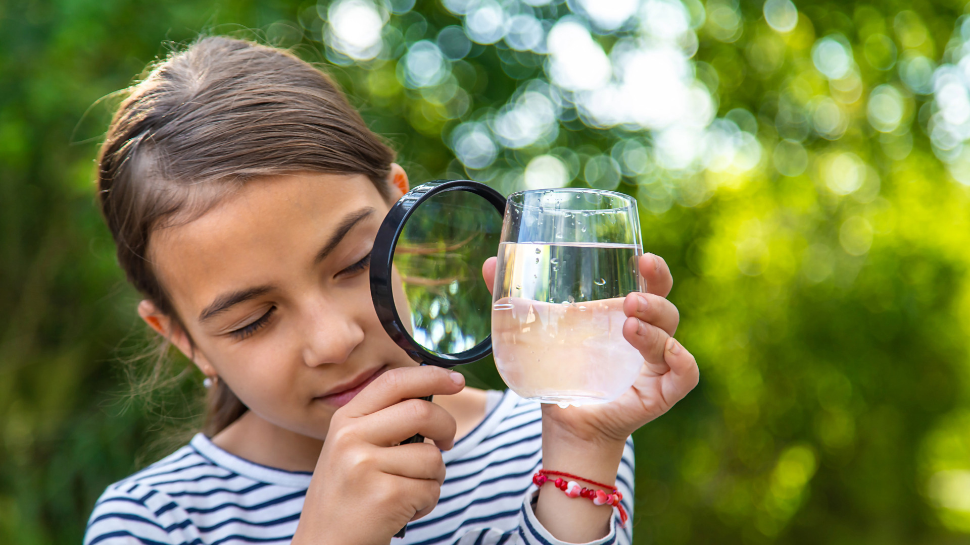 Young girl outdoors, holding a glass of water in one hand and a magnifying glass in the other, examining the water with her eyes closed, surrounded by green blurred foliage.