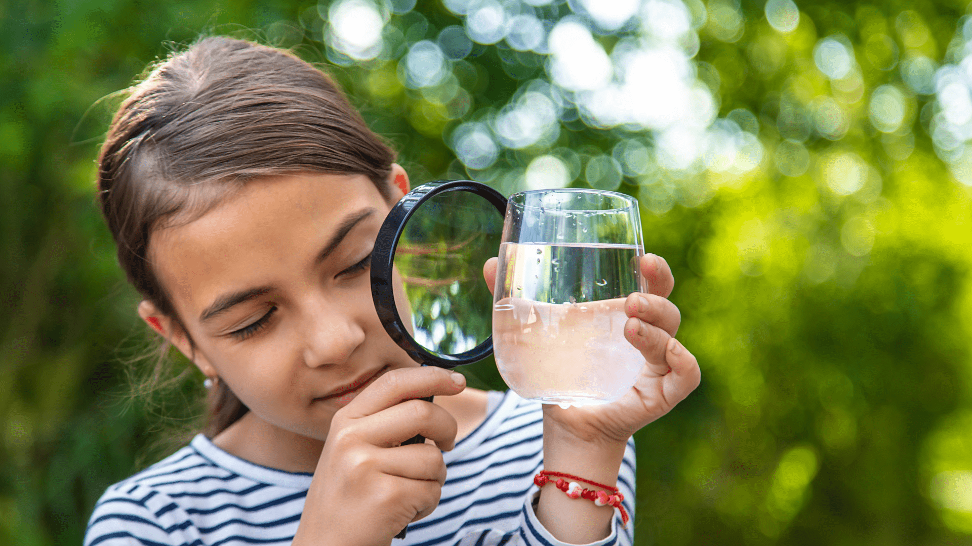 Girl with brown hair holding a glass of water and examining it with a magnifying glass outdoors with green trees background.