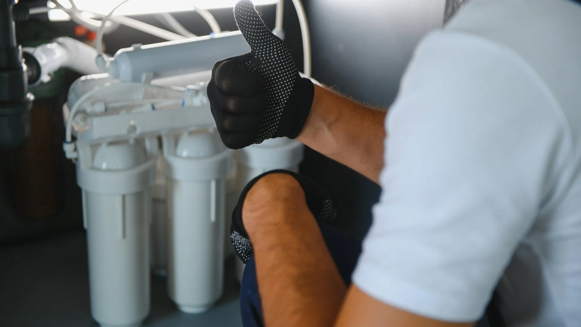 Person working on a water filtration system with three white reverse osmosis filter canisters, wearing black gloves and a white shirt.