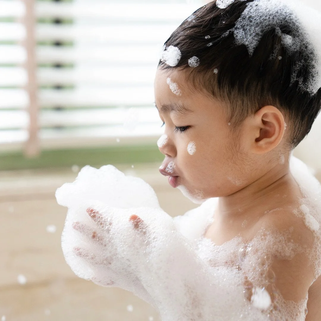 A young boy washing himself with soap, with bubbles on his face, head, and arms, during bath time.