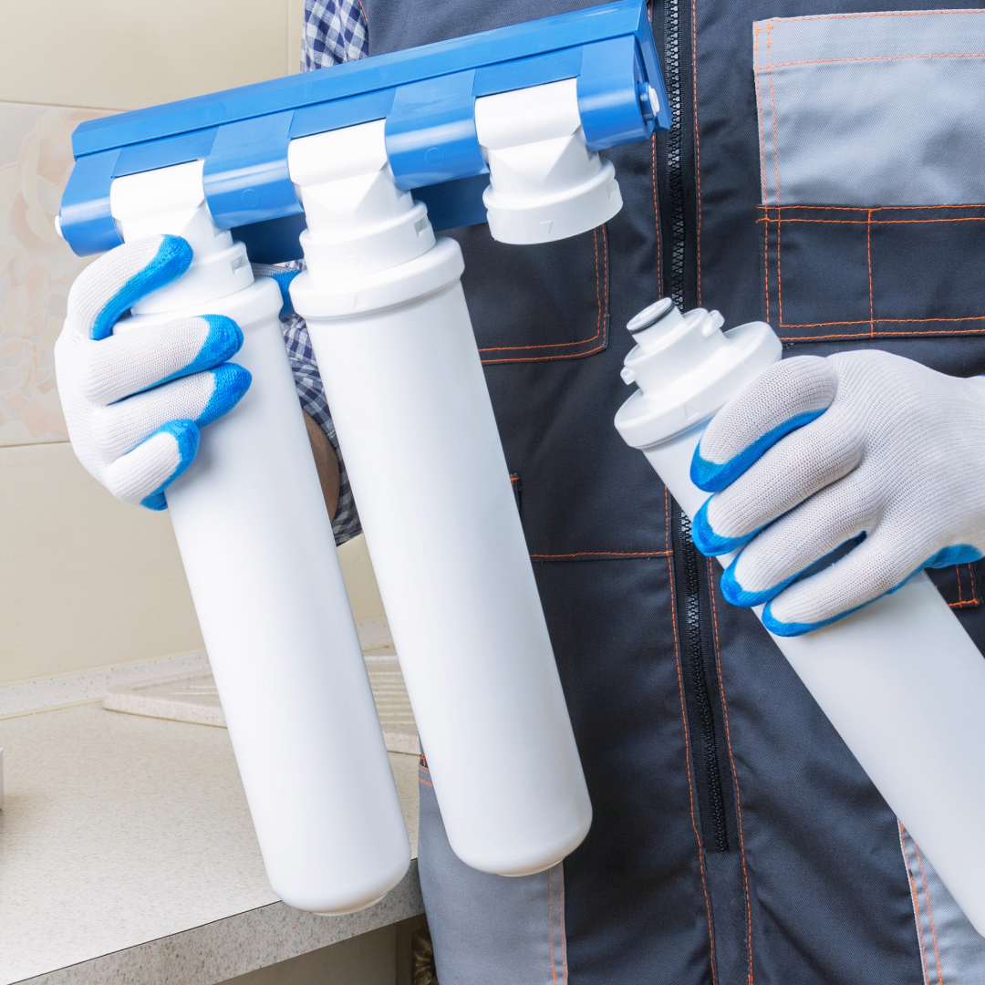 Person holding a water filter replacement cartridge for reverse osmosis system along with a filter housing unit in a kitchen or utility room.