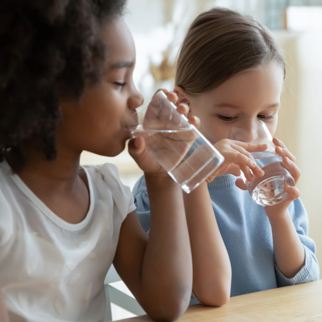 Two young girls sitting at a table, drinking water from clear glasses, with a bright, out-of-focus background.