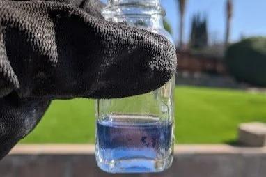 Close-up of a person's nose holding a small clear glass jar with blue water quality testing liquid, outdoors with grass and trees in the background.