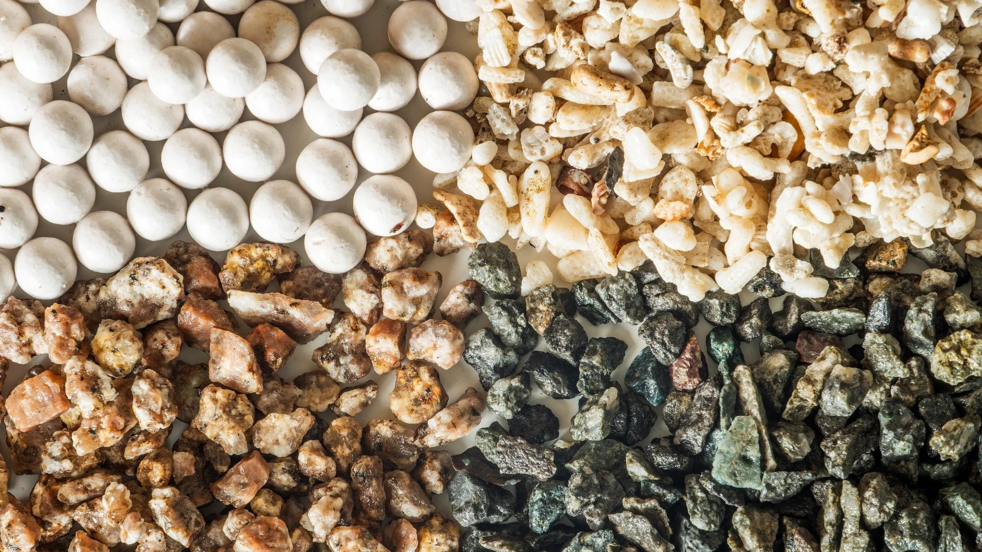 A variety of rocks and minerals arranged in rows, including white round pebbles, small colorful gravel, and darker rough stones.