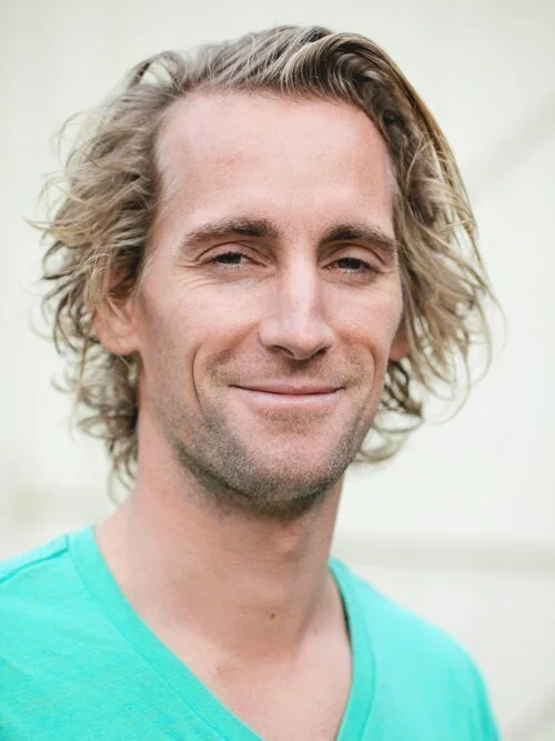A man with shoulder-length wavy blonde hair, fair skin, and a subtle smile, wearing a turquoise T-shirt.