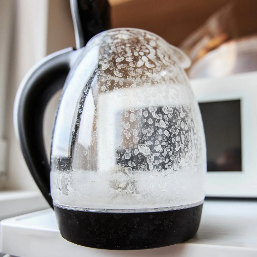 A glass electric kettle with a black handle and base, showing condensation on the inside, sitting on a white surface with a microwave in the background.