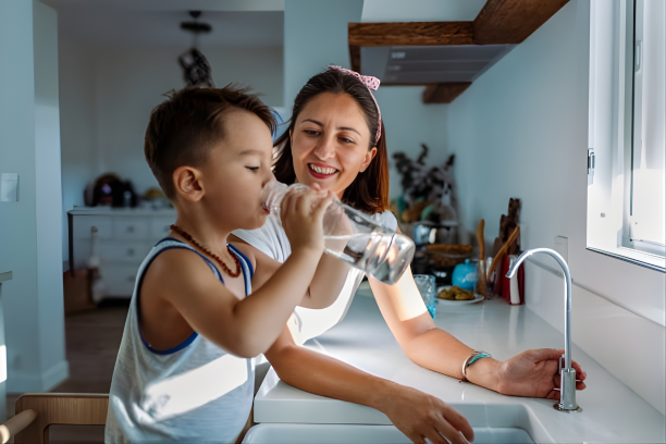A woman helping a young boy drink water from a glass at a kitchen sink.