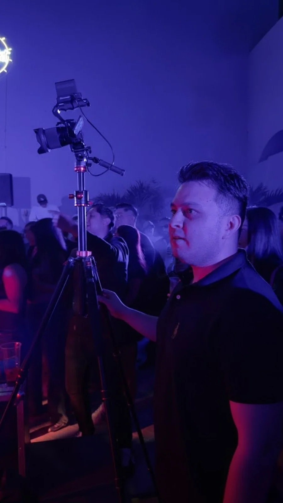 A man holding a camera on a tripod at a crowded indoor event with purple and pink lighting.