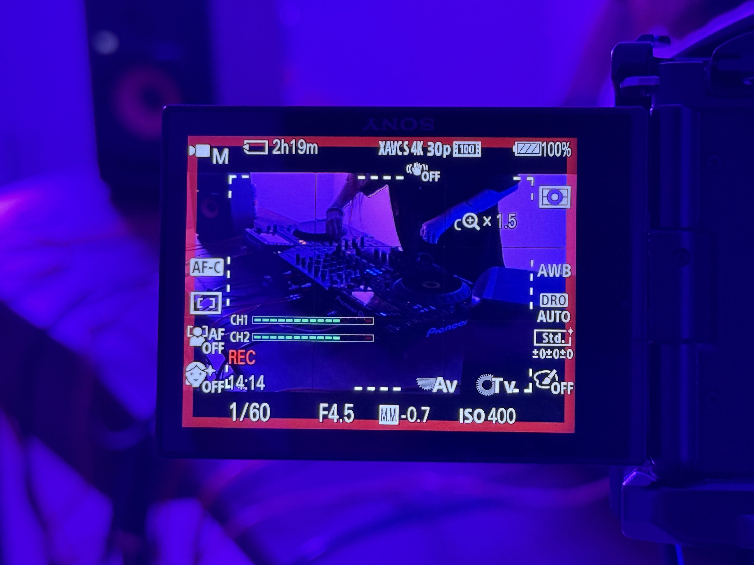 View through a camera screen showing a DJ turning a mixing deck under purple lighting.