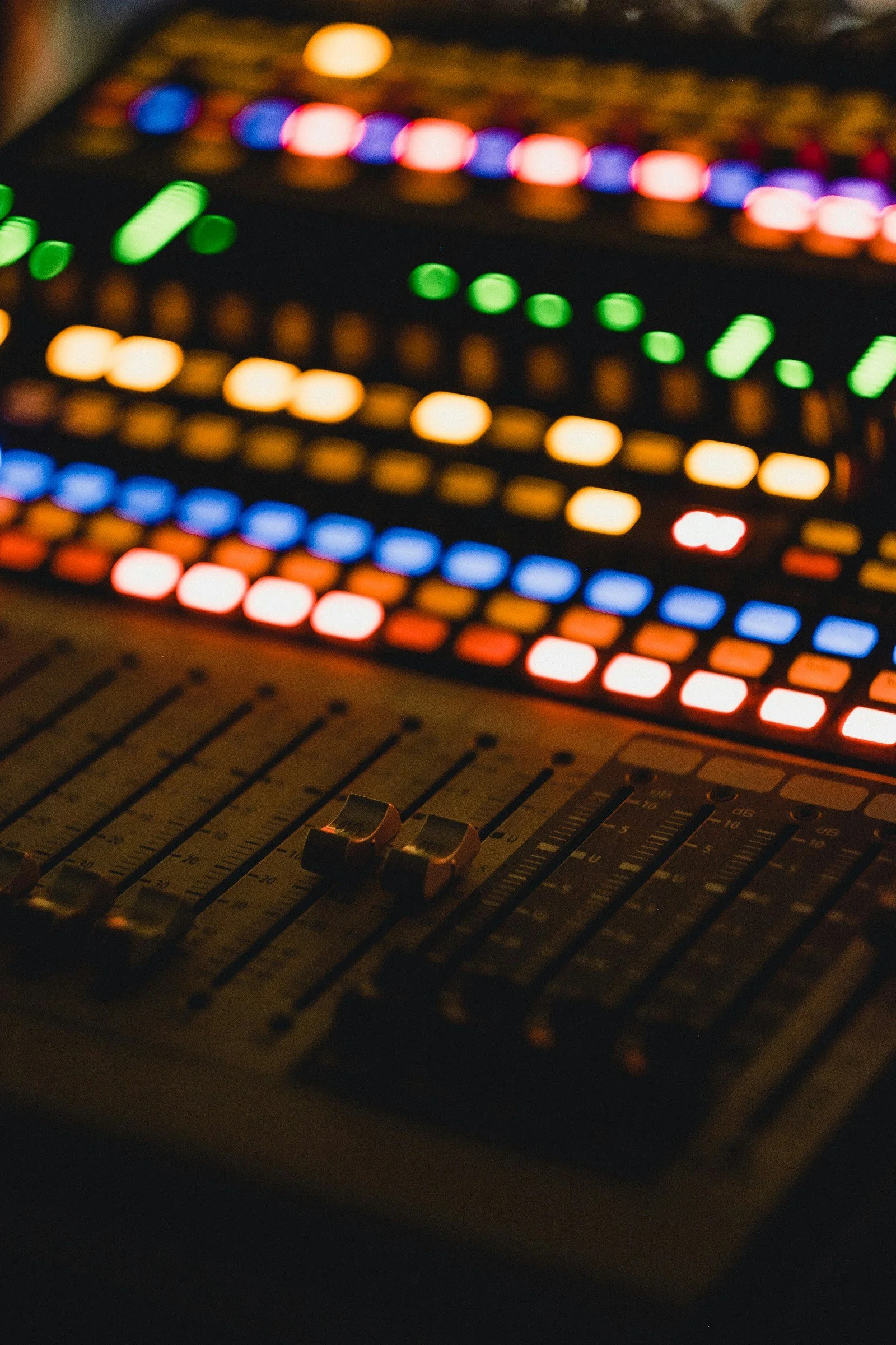 A close-up view of a sound mixing console with sliders and controls, illuminated by colorful LED lights in the background.