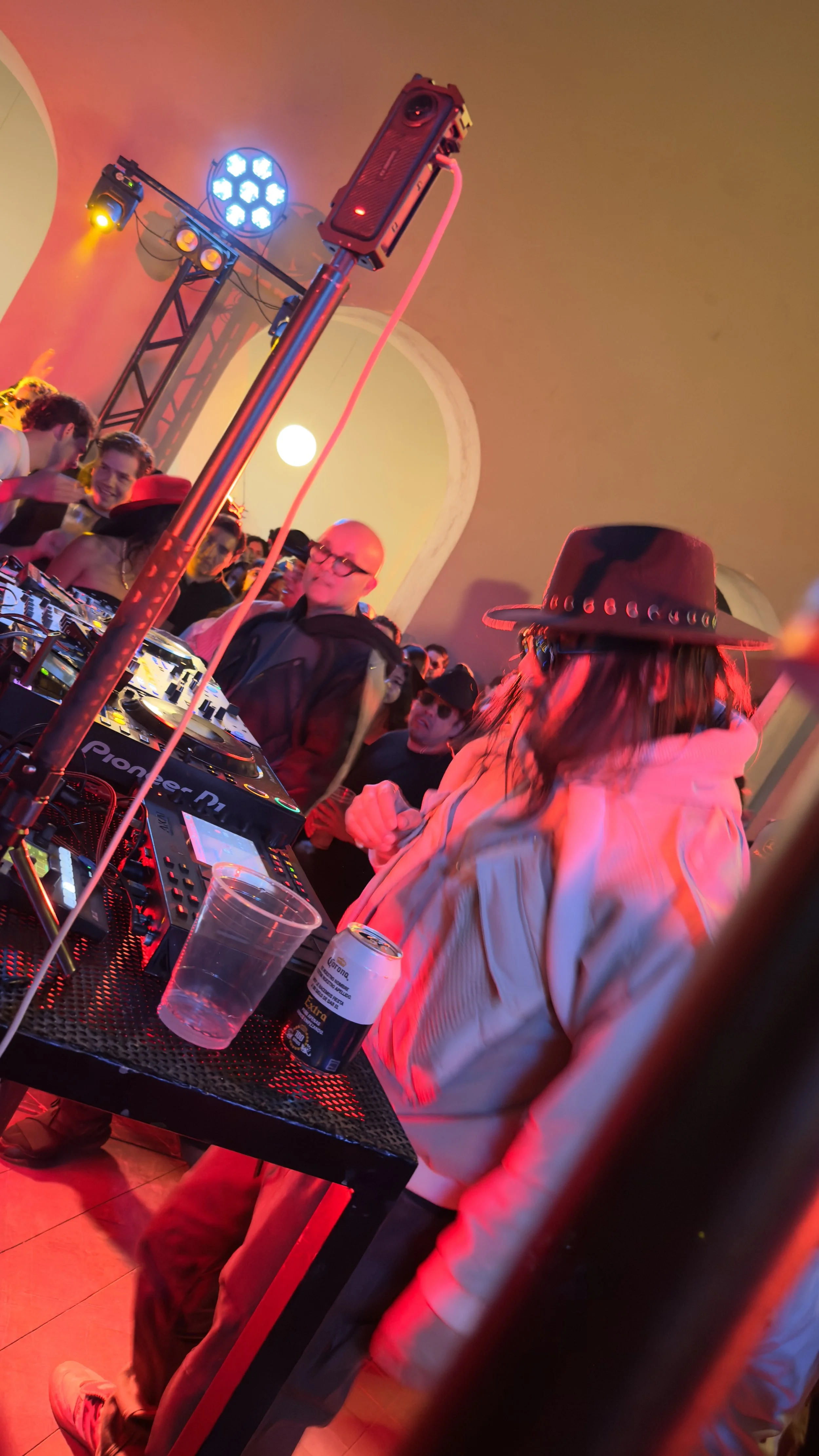 A DJ wearing a cowboy hat and a beige jacket is playing music at a crowded indoor party with colorful lighting. There are people dancing and socializing around the DJ booth, with music equipment visible on the table.