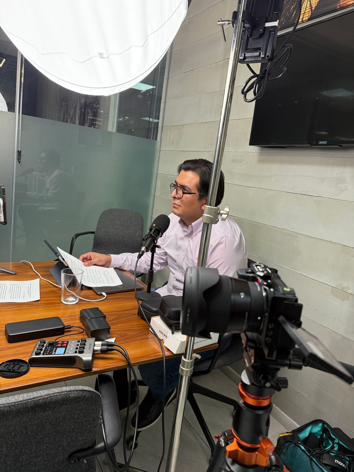 Man wearing glasses sitting at a wooden table, reading from papers, with recording equipment, camera, and lighting setup for a photo or video interview in an office or studio setting.