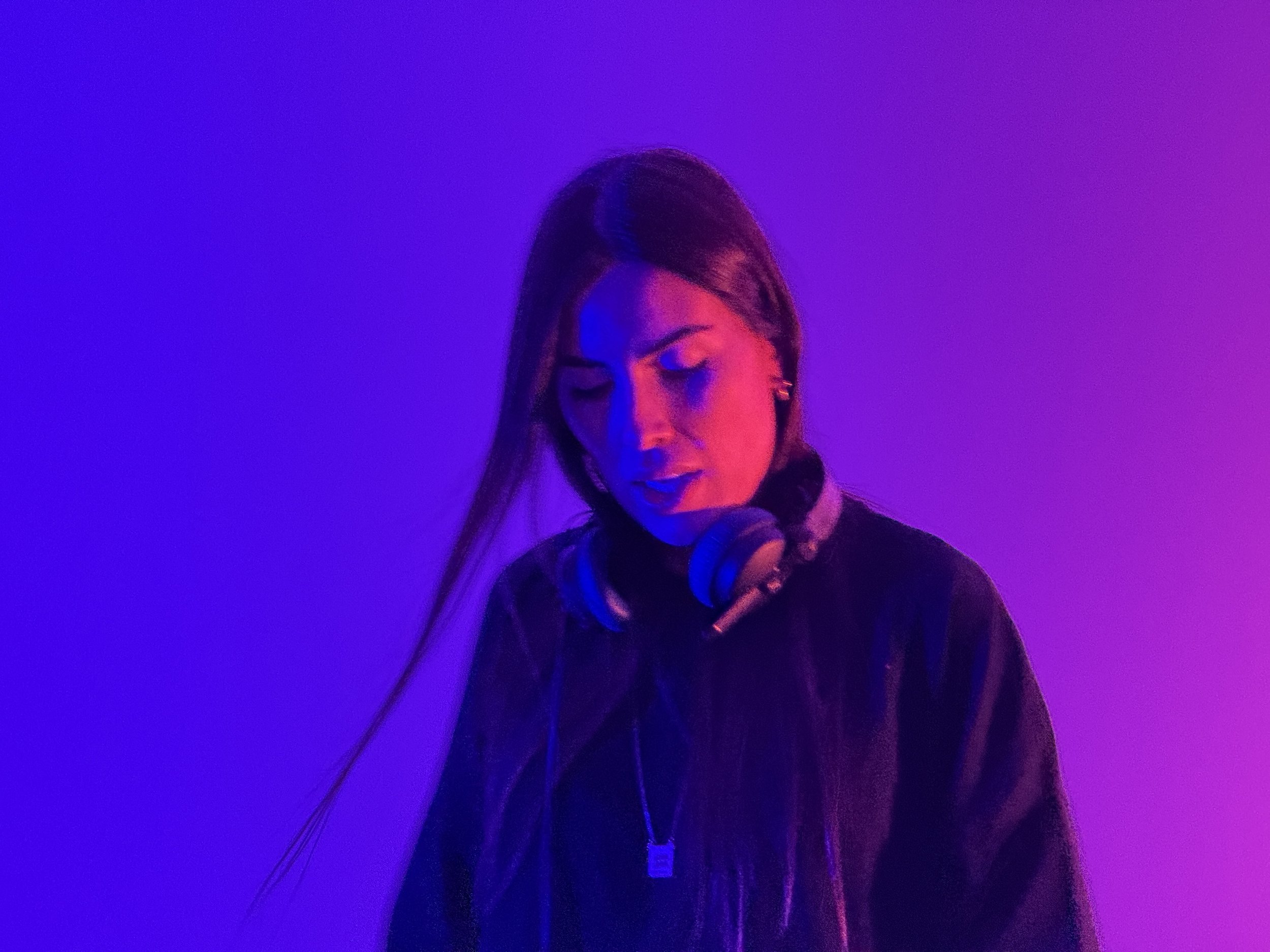 A woman with long dark hair, wearing a black jacket, headphones around her neck, and earrings, is looking down in a room illuminated with purple and pink lighting.