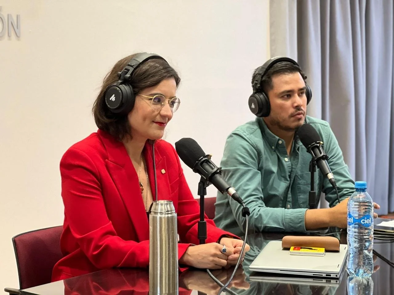 Two people wearing headphones and speaking into microphones at a conference table. The woman on the left is wearing a red blazer and glasses, and the man on the right is wearing a green shirt. There are water bottles, a laptop, and a thermos on the t
